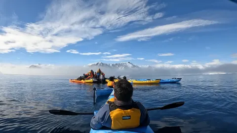 A view from a kayak across calm water towards a group of kayaks and mountains in the mist.