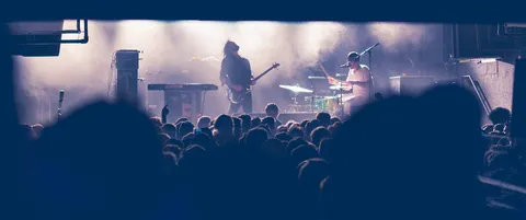 A band on a smoky stage seen over the heads of the crowd, a bassist and a drummer lit from behind